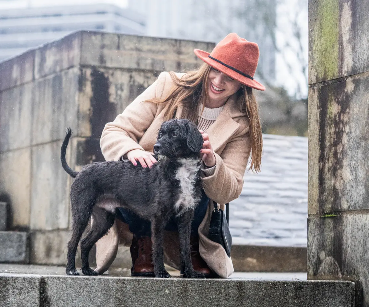 Woman in a red hat kneeling beside her black dog in an urban setting
