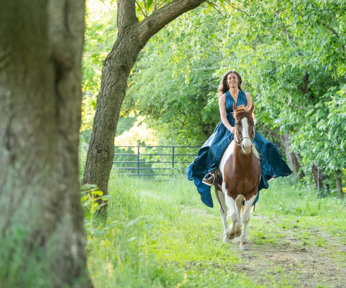 Woman riding a brown and white horse beneath green trees